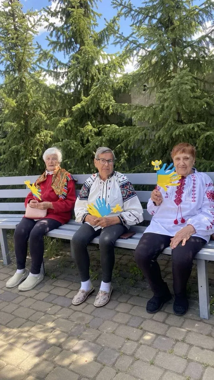 Троє мешканців сидять на лавці серед дерев і проводять час на відкритому подвір’ї.